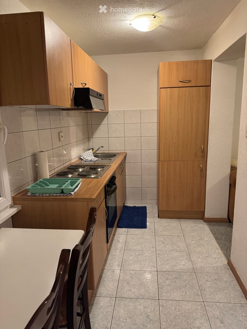 Compact kitchen with wooden cabinets, a refrigerator, an oven, a sink, and a small dining area with a chair.