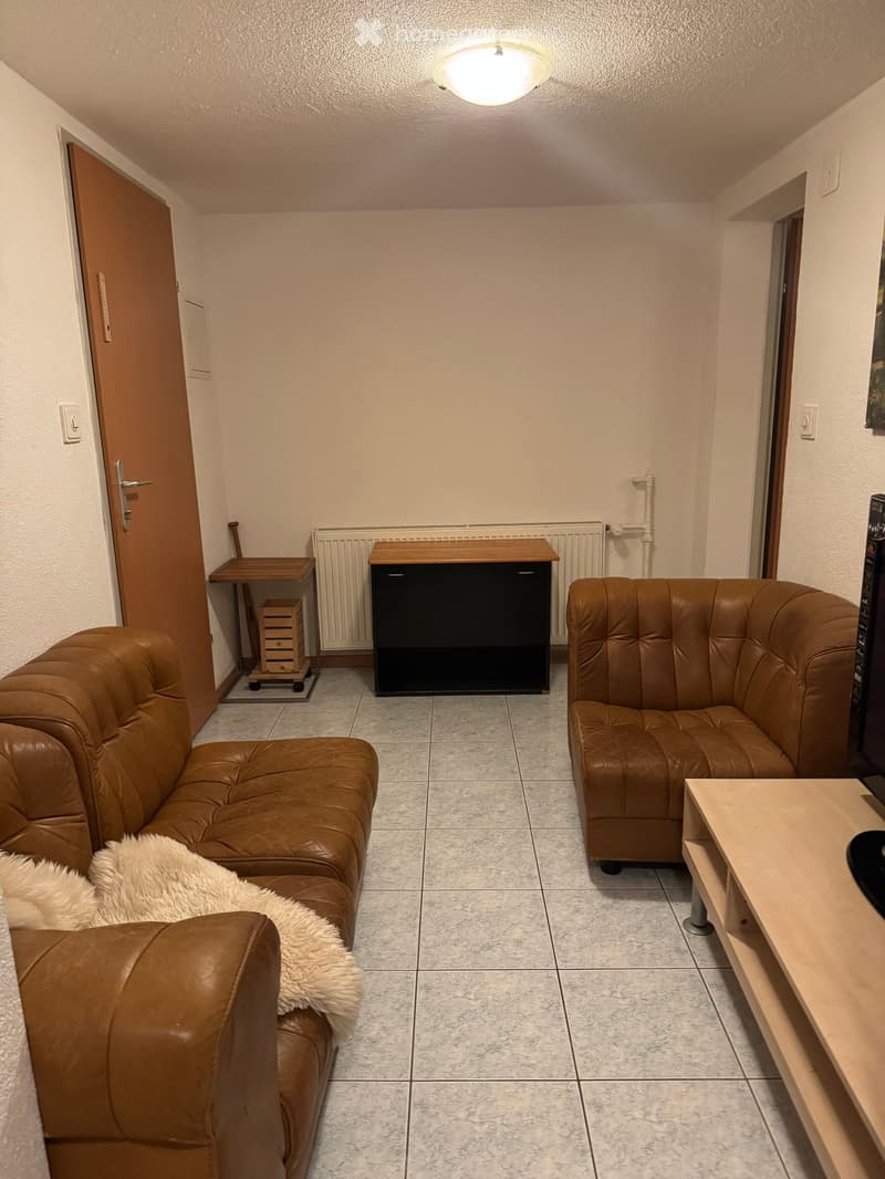 A cozy living room with a brown leather sofa, matching armchair, small wooden table, and a cabinet. The room features tiled flooring and a radiator against the wall.
