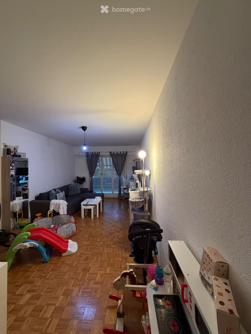 Living room with wooden flooring, a dark-colored sofa, coffee table, baby stroller, toy slide, and kitchen counter with baby items.