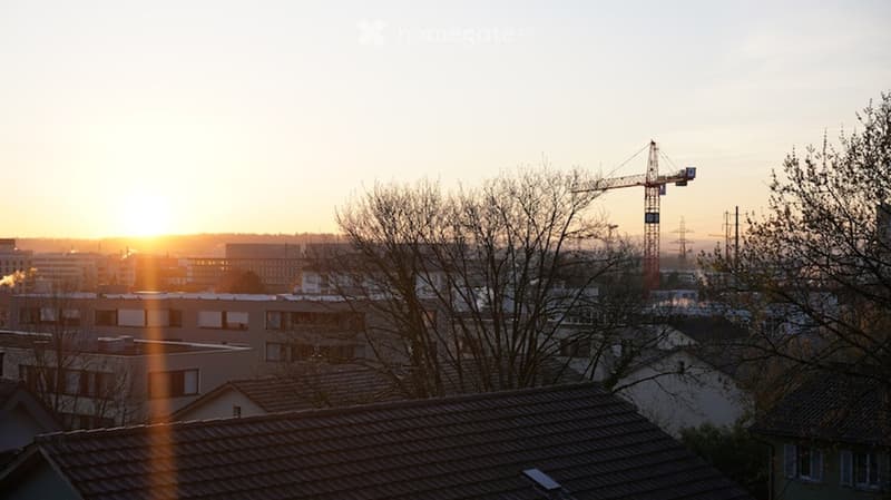 A cityscape view at sunset, showing multiple buildings, rooftops, and a construction crane in the distance.