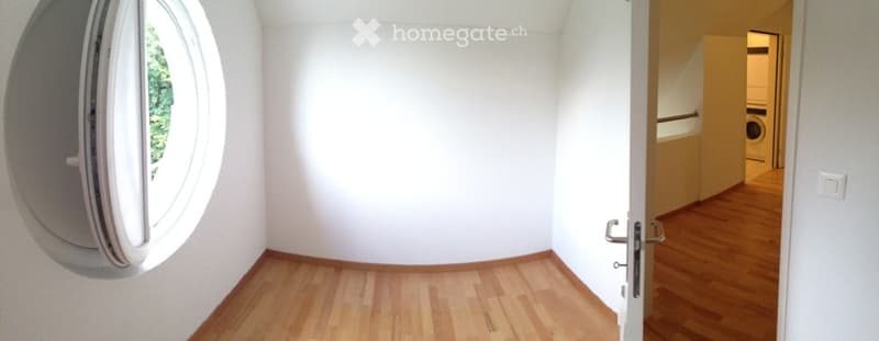 Empty room with wooden flooring, white walls, and a round window. A door leads to a hallway with a washing machine visible in the background.