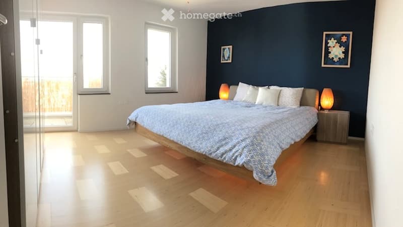 Modern bedroom with wooden flooring, large windows, and a balcony. Features a bed with white pillows and a blue duvet, nightstands with table lamps, and artwork on the walls.