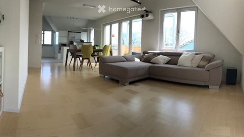 Bright living room with a grey sectional sofa, a dining area with a table and chairs, large windows, and wooden flooring.