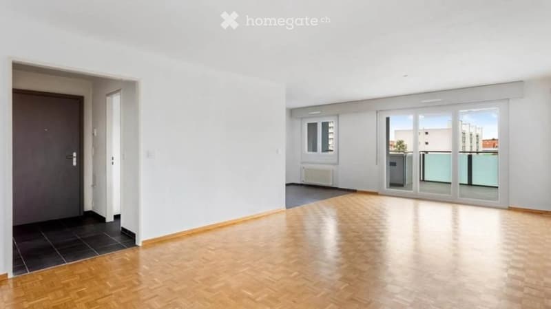 Empty room with wooden flooring, white walls, and large windows leading to a balcony area.