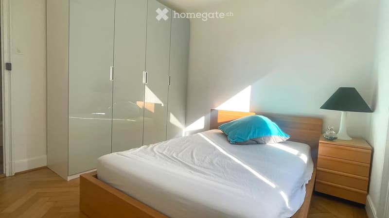 Bright bedroom with a wooden bed, white bedding, a blue pillow, a nightstand with a lamp, and a large mirrored wardrobe.