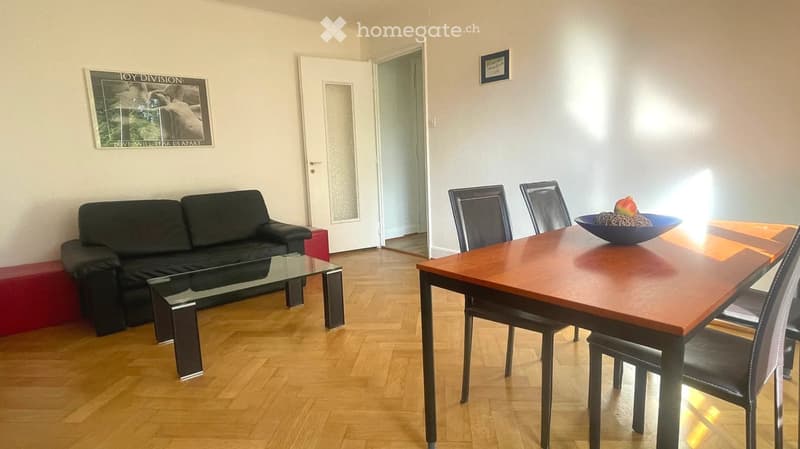 A modern living and dining area with a black sofa, glass coffee table, wooden dining table, and four chairs. A bowl with fruit sits on the dining table. The room features light-colored walls, wooden flooring, and a framed poster on the wall.