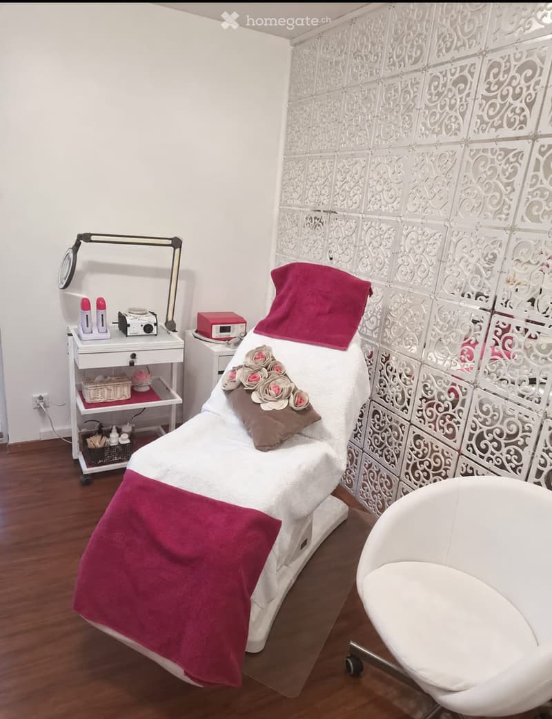 A well-organized massage or treatment room with a massage table, a white chair, a small cabinet with various items, and a decorative wall with a patterned design.