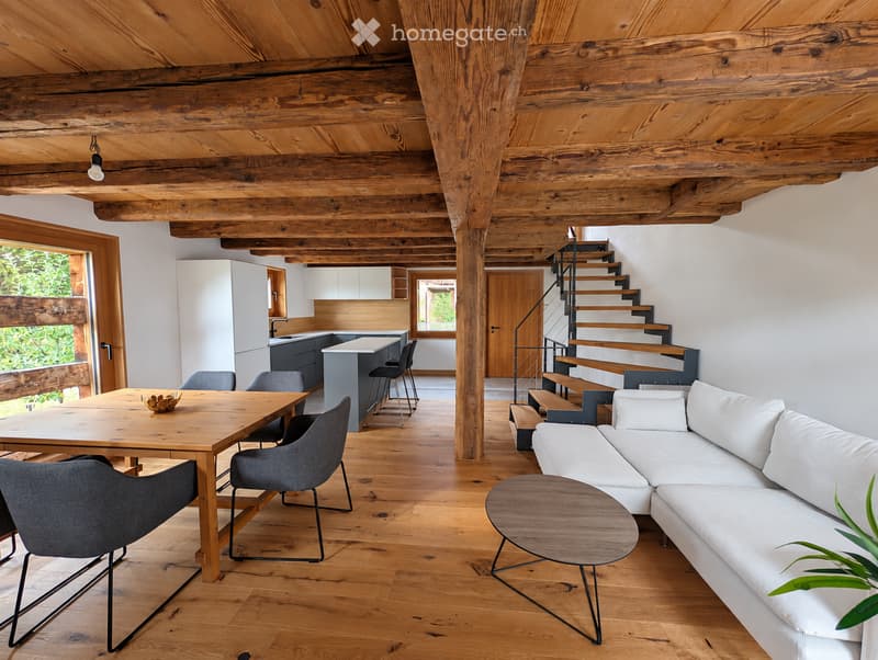 Open-concept living and dining area with exposed wooden beams, hardwood flooring, white walls, and modern furniture including a white sectional sofa, wooden coffee table, dining table with chairs, and a kitchen island with bar stools.