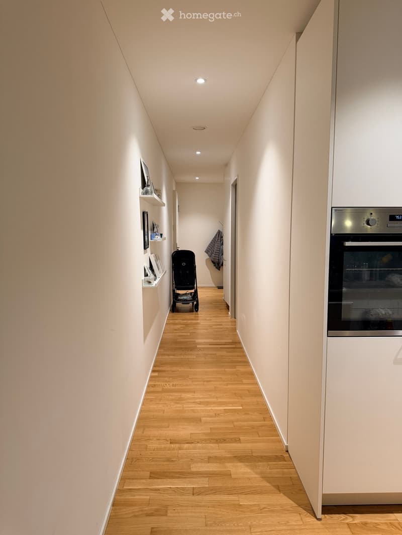 A narrow hallway with light wooden flooring, white walls, and built-in shelves on one side. A stroller is positioned in the center, and a modern oven is visible on the right side.