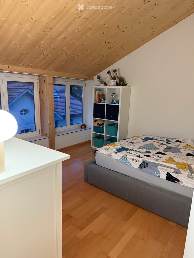 Cozy attic bedroom with wooden slanted ceiling, hardwood floor, large windows, bed with colorful bedding, shelf with books and decorative items, and a modern lamp on the left side.