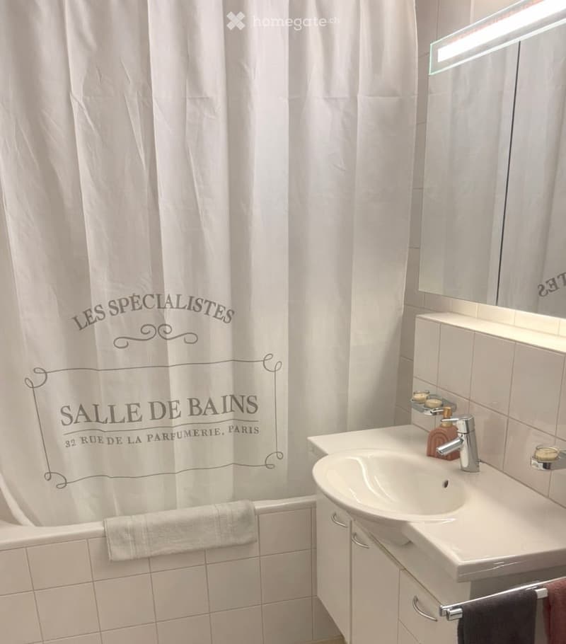 Modern bathroom with a white bathtub, shower curtain, sink, and mirror. The space features white tiles and a sleek design.