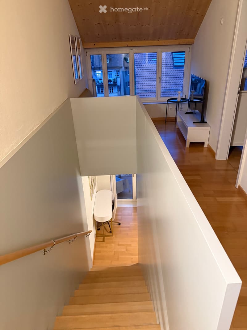 Modern staircase with wooden steps and a wooden handrail, leading to an open landing area with views of adjacent rooms and a balcony.