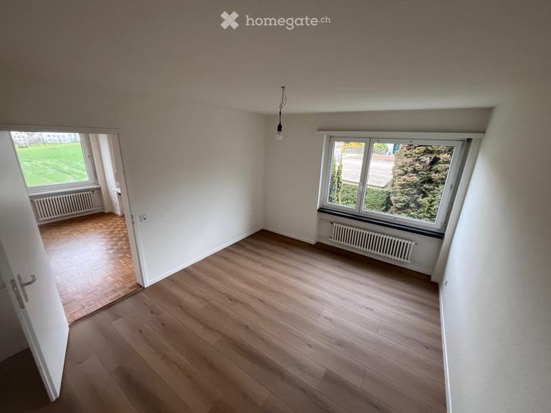 Empty room with wooden flooring, white walls, and large windows offering views of greenery. Radiators are visible beneath the windows, and a pendant light hangs from the ceiling.
