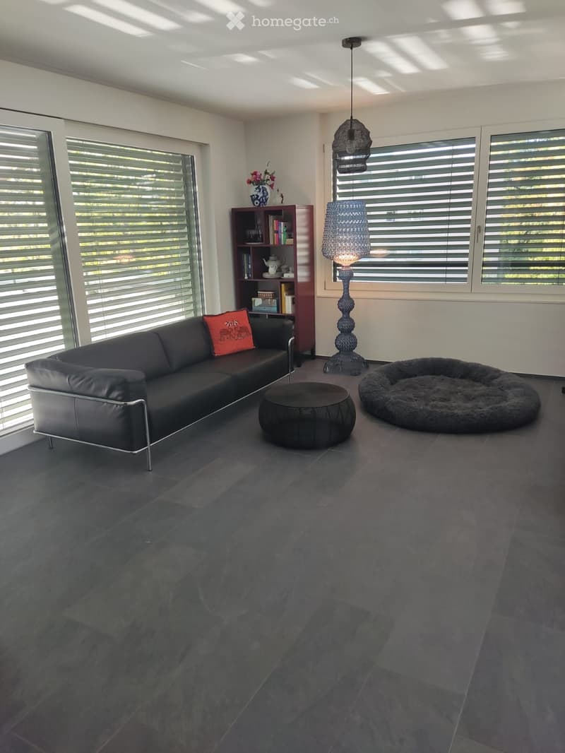 Modern living room with dark grey tiles, large windows with blinds, a black leather sofa with red throw pillow, a round black ottoman, a tall decorative floor lamp, a bookshelf with books and decorative items, and a plush dog bed.
