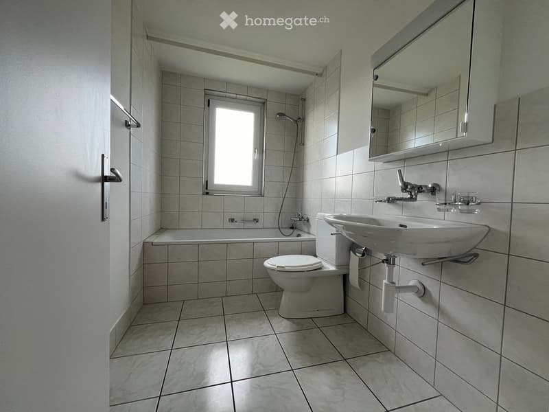 Modern bathroom with tiled walls and floor, featuring a toilet, sink, bathtub, and mirror above the sink.