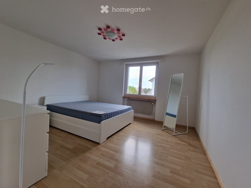 Bright bedroom with wooden flooring, white walls, a large window, a bed with blue bedding, a standing mirror, and a floor lamp.