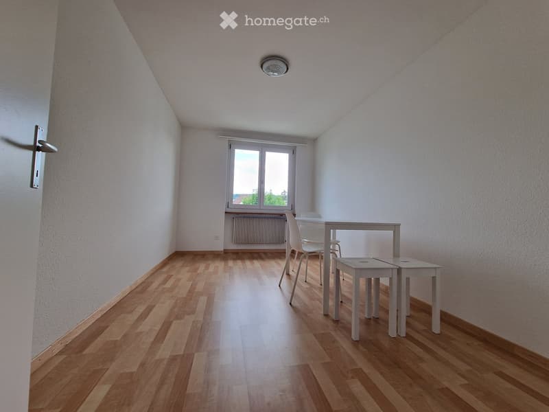 Bright, unfurnished room with wooden flooring, white walls, and a large window. Includes a small table, chairs, and stools.