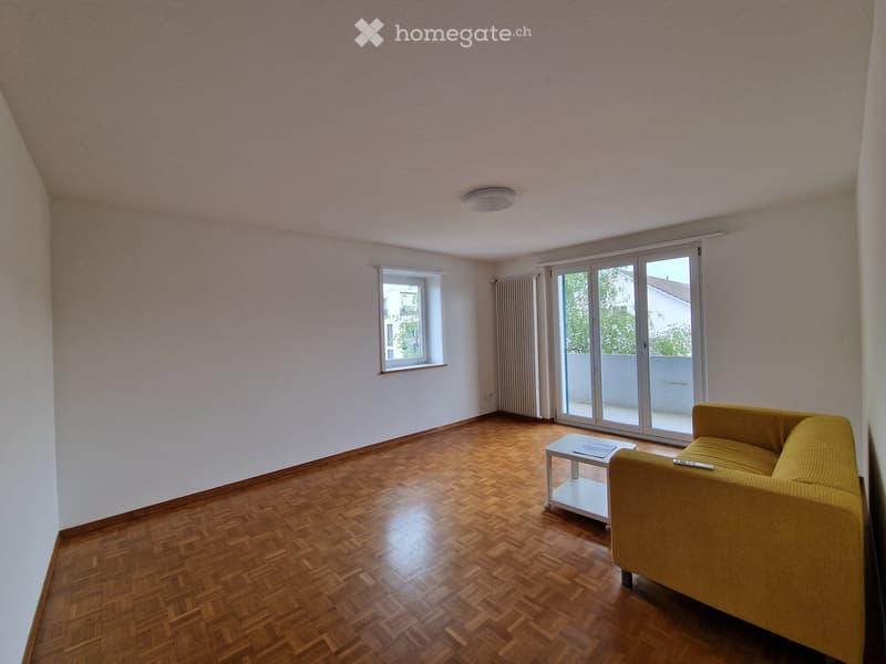 Bright living room with wooden parquet flooring, large sliding glass doors leading to a balcony, a yellow sofa, and a white coffee table.