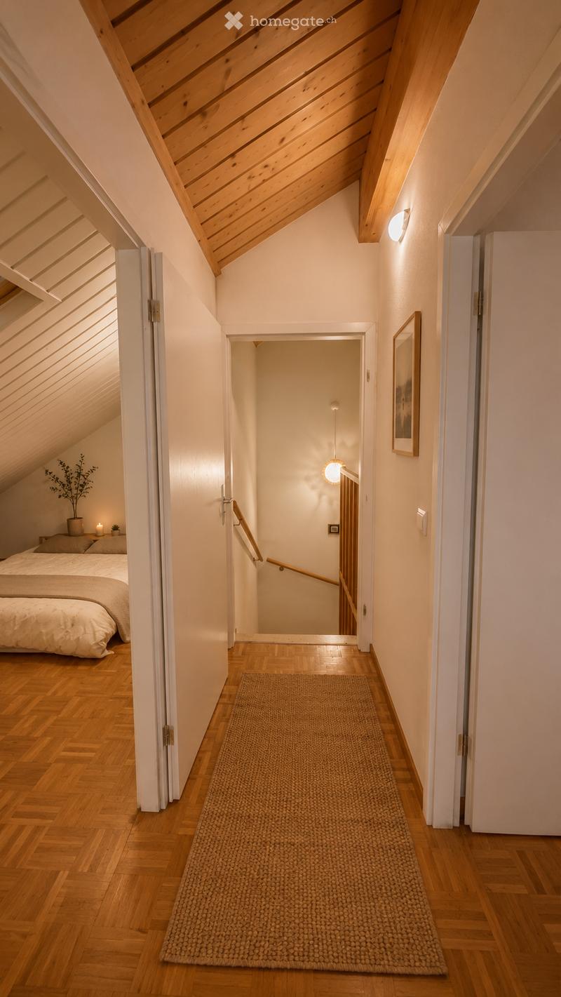 A narrow hallway with wooden flooring, a woven runner, and a staircase leading upwards. The hallway features white walls, a slanted wooden ceiling, and a small bedroom visible to the left with a bed and nightstand.