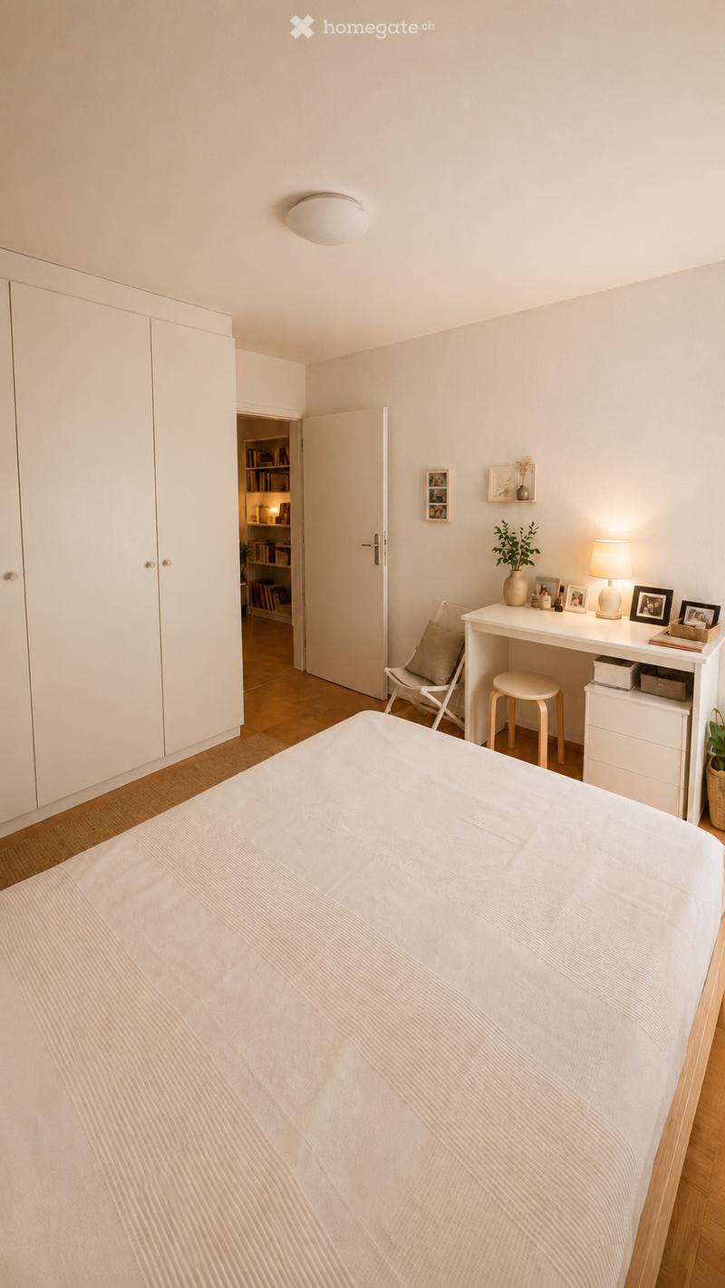 A modern, well-lit bedroom with a neatly made bed, a white desk with a chair, a small stool, a built-in wardrobe, and a small open shelving unit visible through an open door.