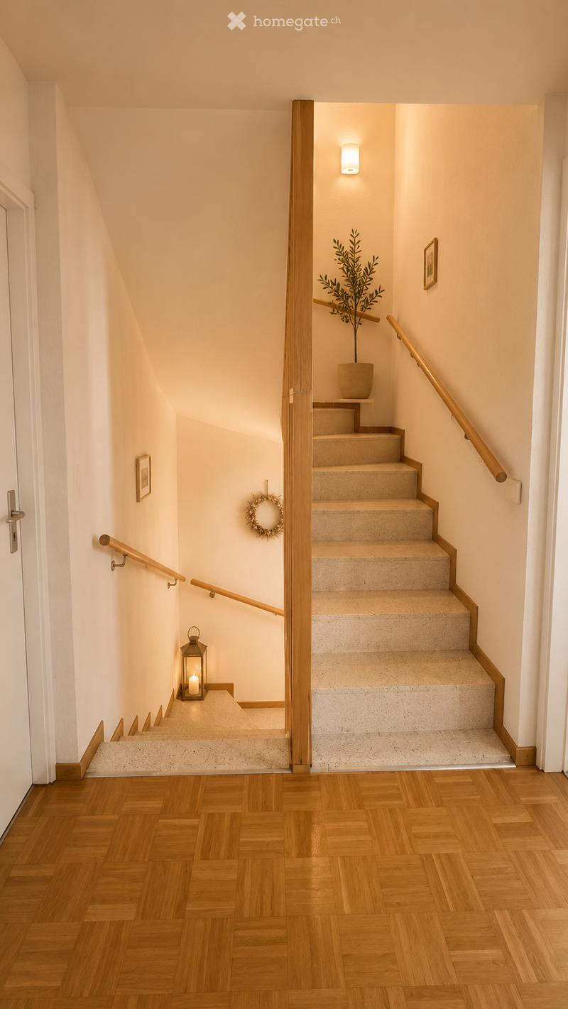 Spacious hallway with wooden flooring, staircase leading upwards, wooden railings, wall-mounted light fixture, small lantern with candle, framed picture, potted plant, and decorative wreath.