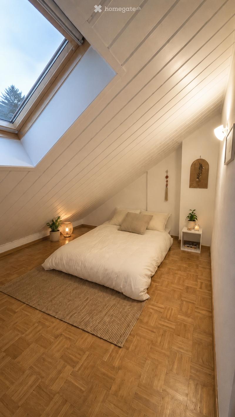 Cozy attic bedroom with wooden herringbone flooring, slanted ceilings, skylight windows, a neatly made bed with pillows and a duvet, a nightstand with a lamp, and a small plant on a side table.