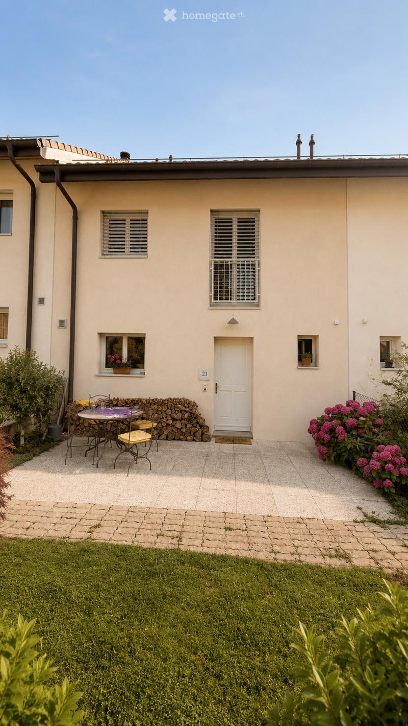 A well-maintained garden with a neatly trimmed lawn, a patio area with a table and chairs, a stack of firewood, and a white front door with a small doormat. The patio is surrounded by colorful flowers and shrubs.