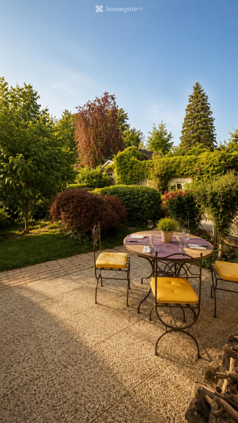 A well-maintained garden with a round outdoor table and four chairs, set on a stone patio surrounded by lush greenery and trees.