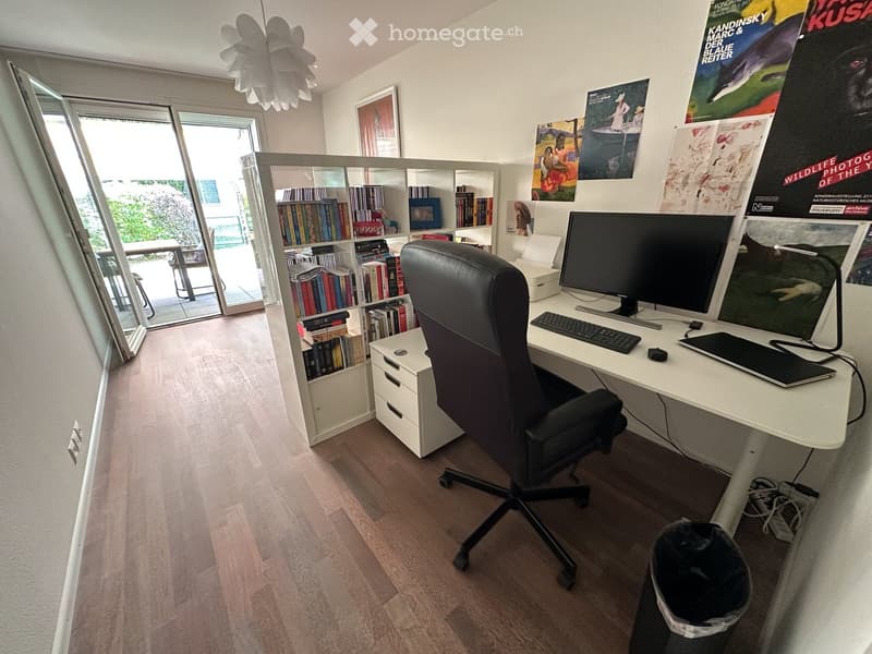 Modern office space with a large desk, ergonomic chair, laptop, keyboard, and a bookshelf filled with books. Large windows provide natural light, and the room features a stylish lamp and various posters on the walls.