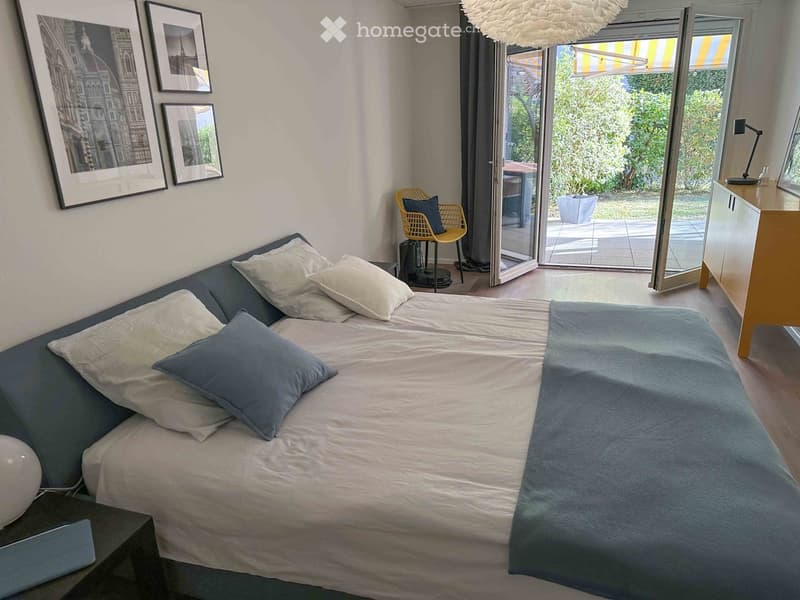 A modern, well-lit bedroom with a large bed, pillows, a nightstand, a small chair, a table, a cabinet, and picture frames on the wall. The room features large windows with a view of a garden.
