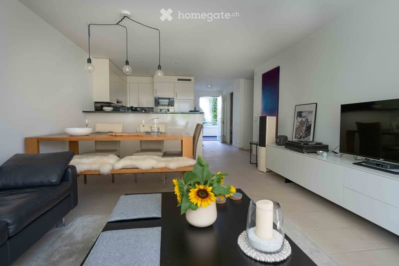 Modern living room with a black leather sofa, coffee table, dining area with wooden table and chairs, white cabinets, and a TV stand with a large screen.