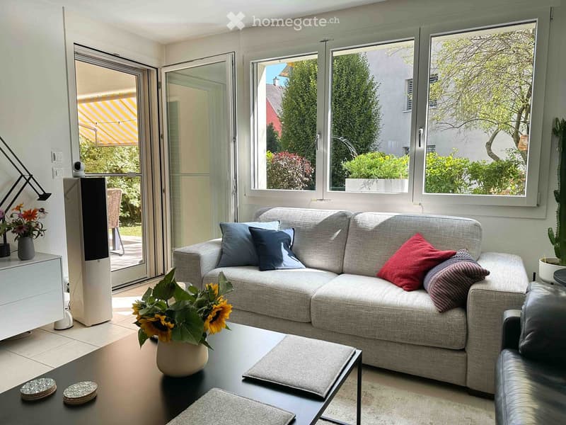 Modern living room with large windows, grey sectional sofa, coffee table, ottoman, armchair, table lamp, and decorative vases.