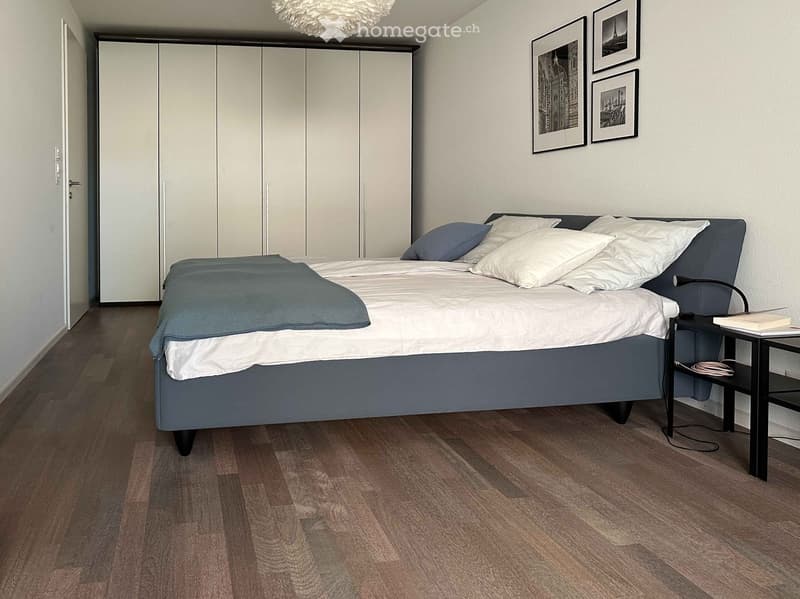 Modern bedroom with a large bed, nightstand, lamp, wardrobe, and picture frames on the wall.