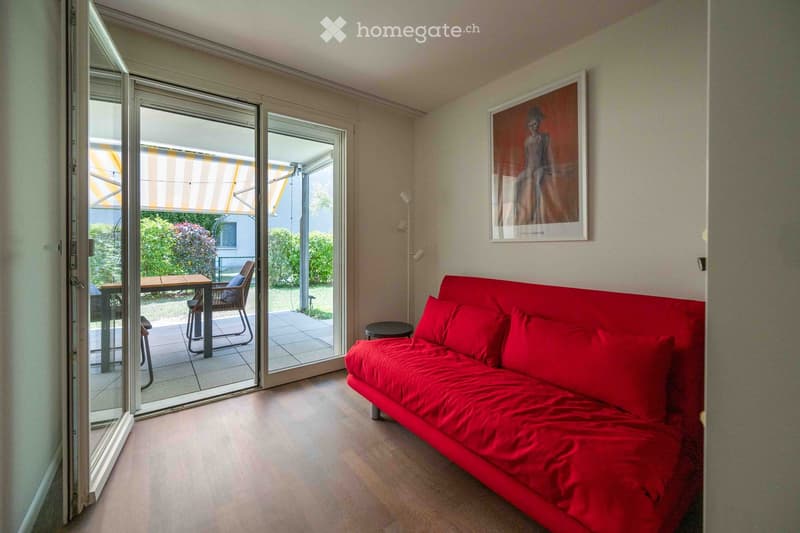 A modern living room with a red sofa, a small table and chairs, a stool, and a picture frame on the wall. Large glass doors open to a balcony with outdoor furniture.