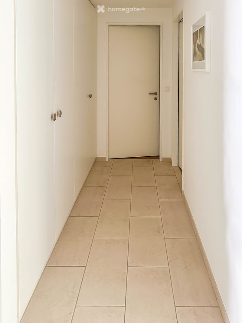 A clean, minimalist hallway with white walls, beige tiled flooring, and a closed door at the end. A framed picture hangs on the right wall.