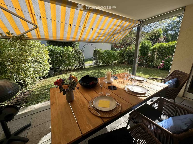 Outdoor dining area with a wooden table set for a meal, surrounded by greenery and a covered patio with striped awning.
