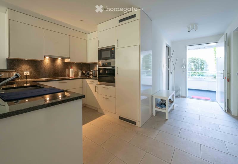 Modern kitchen with white cabinets, stainless steel appliances, and a black countertop. Features a sink, microwave, oven, and a small white stool near the entrance.