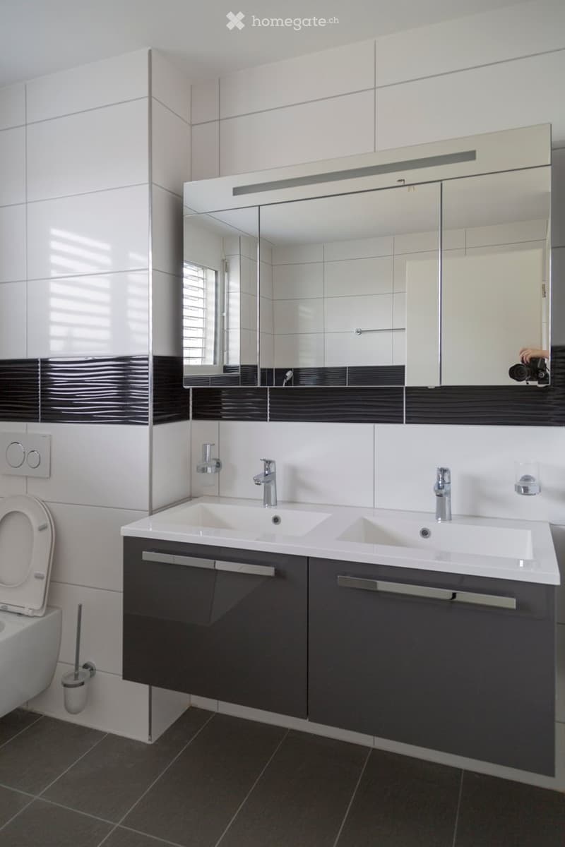 Modern bathroom with white tiles, dark grey cabinetry, double sink, large mirror, and a toilet.