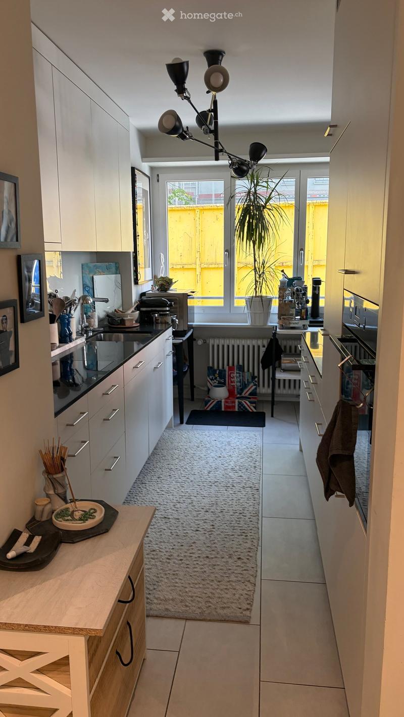 Modern kitchen with white cabinets, black countertops, and a small dining area. Features include a sink, microwave, coffee maker, and a potted plant near the window.