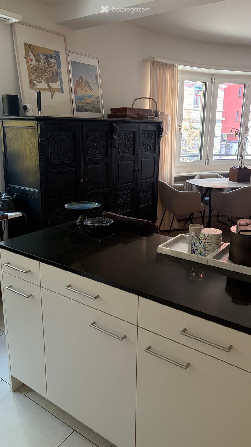 Modern kitchen with white cabinets, black countertop, and various decorative items including a teapot, bowls, and picture frames. There is a dining area with a table and chairs visible in the background.