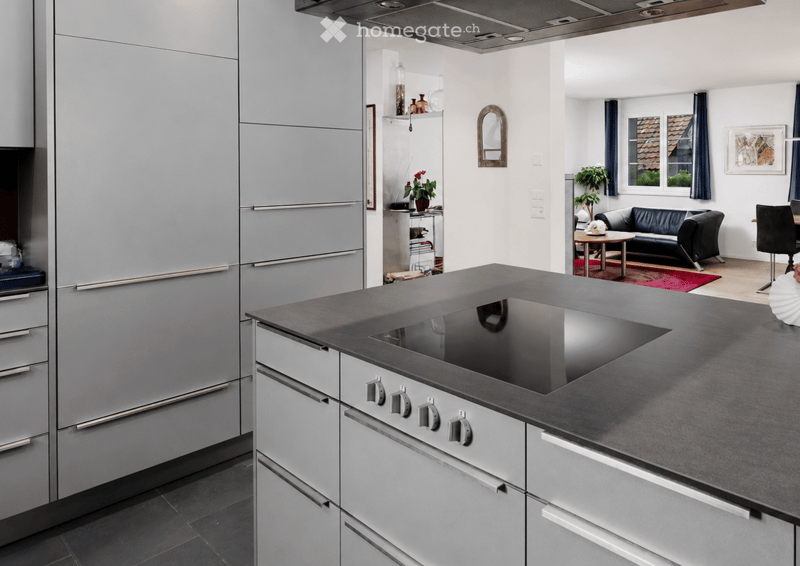 Modern kitchen with stainless steel appliances, grey countertops, and a living area with a black sofa and small table.