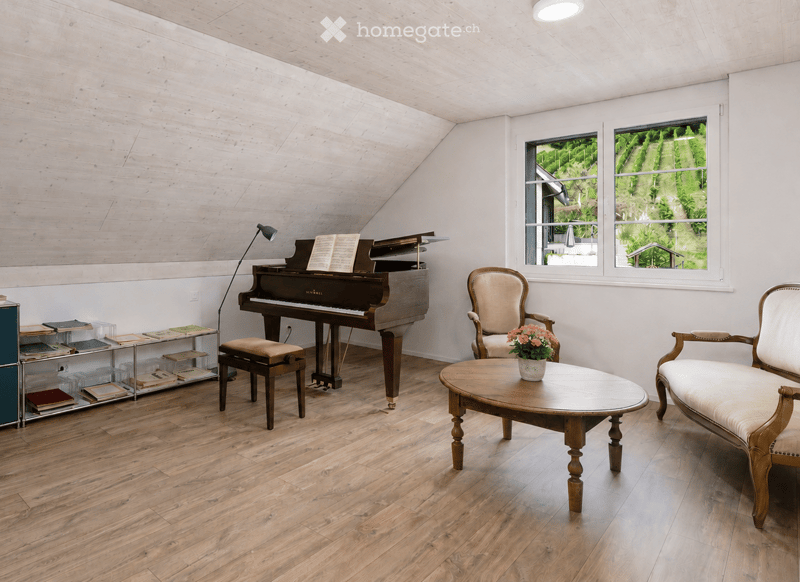 Bright room with wooden floors and slanted wooden ceiling, featuring a grand piano, a small bench, two armchairs, a coffee table with a flower vase, a bookshelf, and a floor lamp. Large windows offer a scenic view of greenery.
