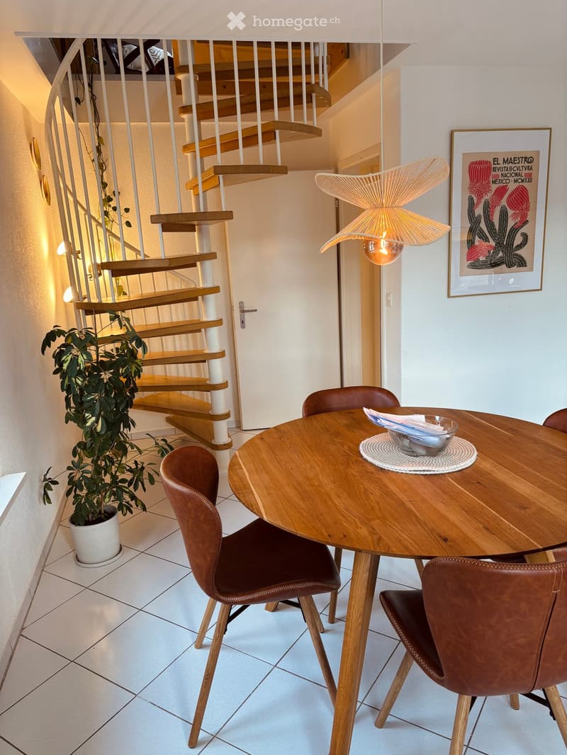 A modern dining area with a round wooden table, four brown leather chairs, a hanging lamp, a potted plant, and a framed picture on the wall. A staircase with wooden steps and white railings is visible in the background.