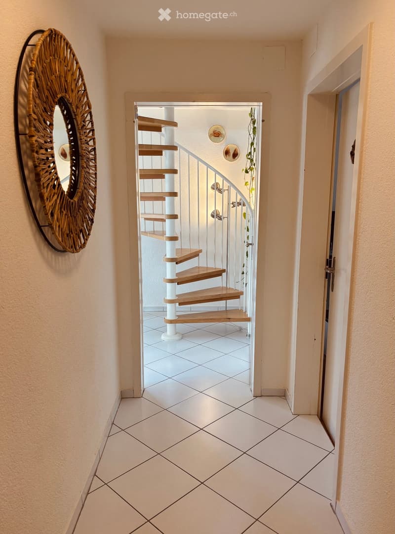 Hallway with white tiled flooring, beige walls, and a decorative woven mirror on the left. A white and wooden spiral staircase leads upwards, with a small plant visible in the background.
