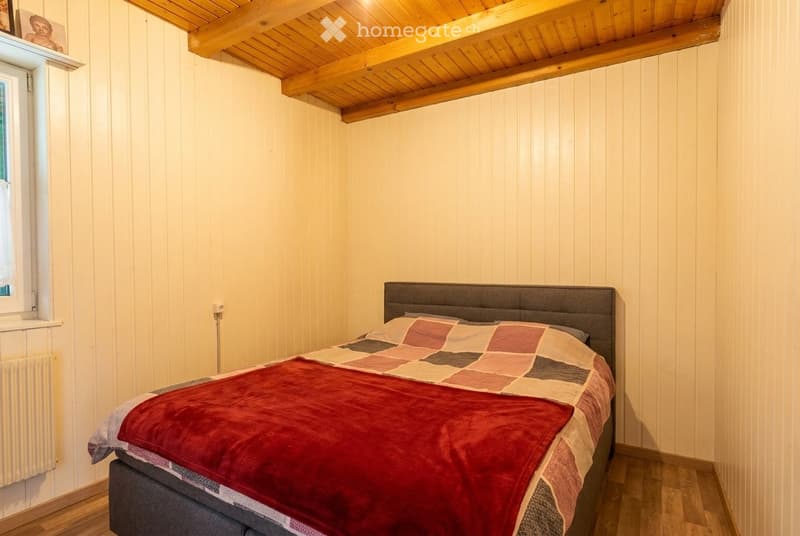 Cozy bedroom with wooden walls, exposed wooden ceiling beams, and a neatly made bed with a colorful patchwork quilt and red throw.