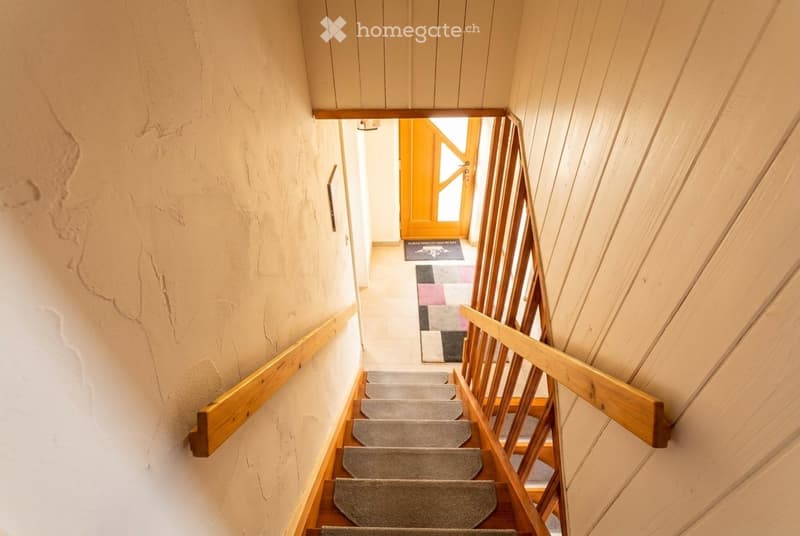 Wooden staircase with gray carpeted steps and wooden railings, leading to a doorway with a colorful rug in the background.