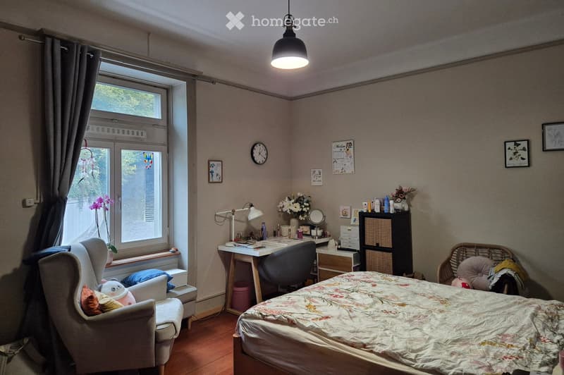 Cozy bedroom with a bed, desk, chair, and decorative items. Features a large window with curtains, a hanging pendant light, and a wicker chair with pillows.