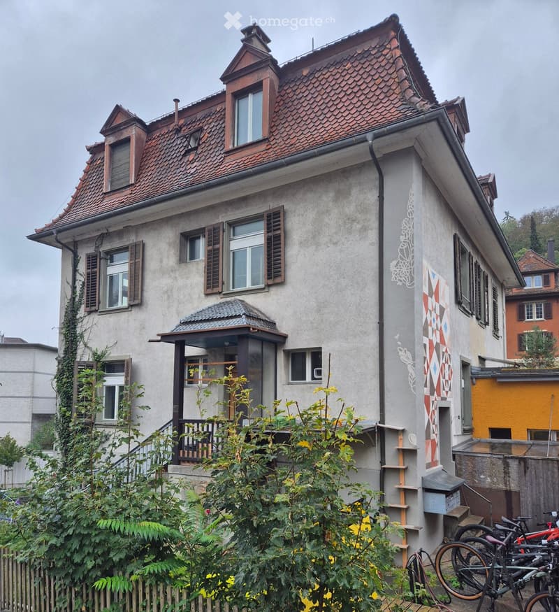 Two-story house with a red-tiled roof, multiple windows with shutters, and a small balcony. The facade features decorative wall art and a small garden with bicycles parked nearby.