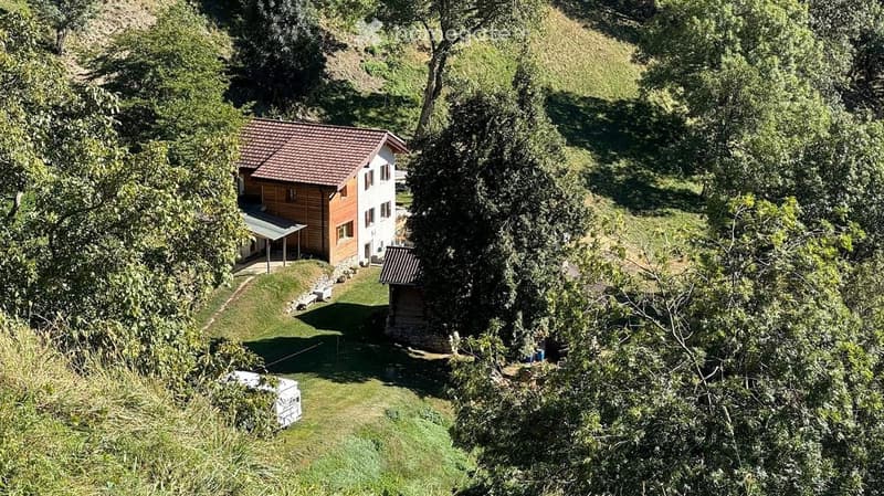 A two-story house with a red-tiled roof and white walls, surrounded by lush greenery and trees. The house features a covered patio area and is situated on a raised ground floor with a small garden and additional structures nearby.