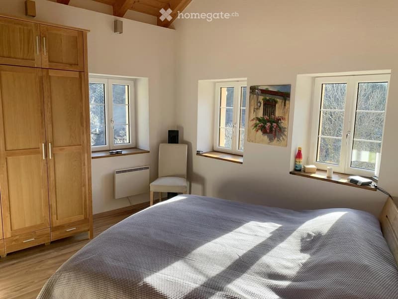 Bright bedroom with wooden floor, large windows, bed with blue bedding, wooden wardrobe, chair, and a painting on the wall.